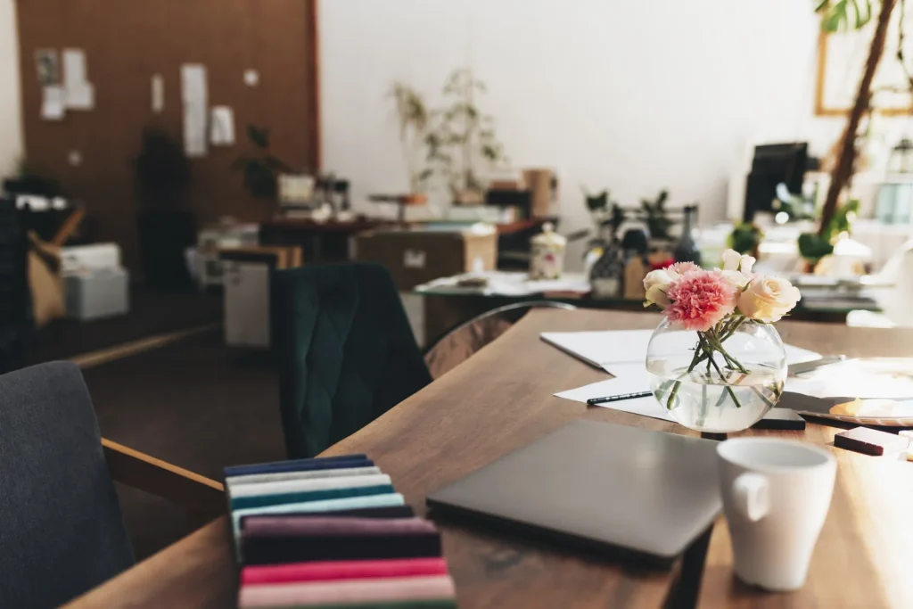 A bright shared workspace table with a laptop, notebook, fabric samples and fresh flowers